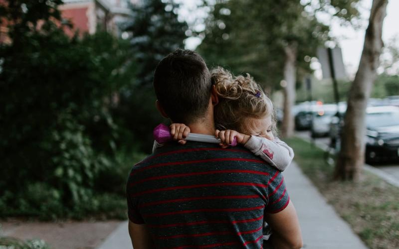 Close to the center of everything tired little girl rests her head on her father's shoulder as he carries her down the city sidewalk